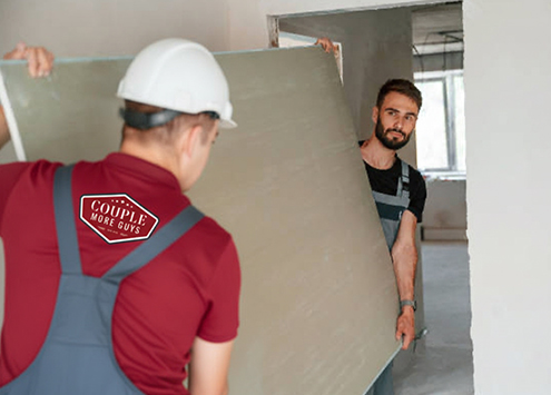 Two construction workers carry a large drywall panel inside an unfinished room. One wears a white hard hat and a shirt with a “Couple More Guys” logo on the back. Both are dressed in work overalls.