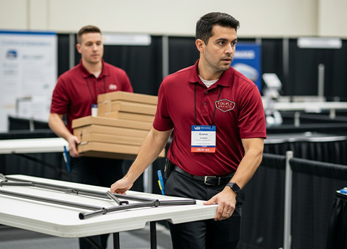 Two men in red polo shirts and name badges are setting up tables at an event. One carries stacked boxes, while the other arranges a table frame, both focused on their tasks in a conference room setting.