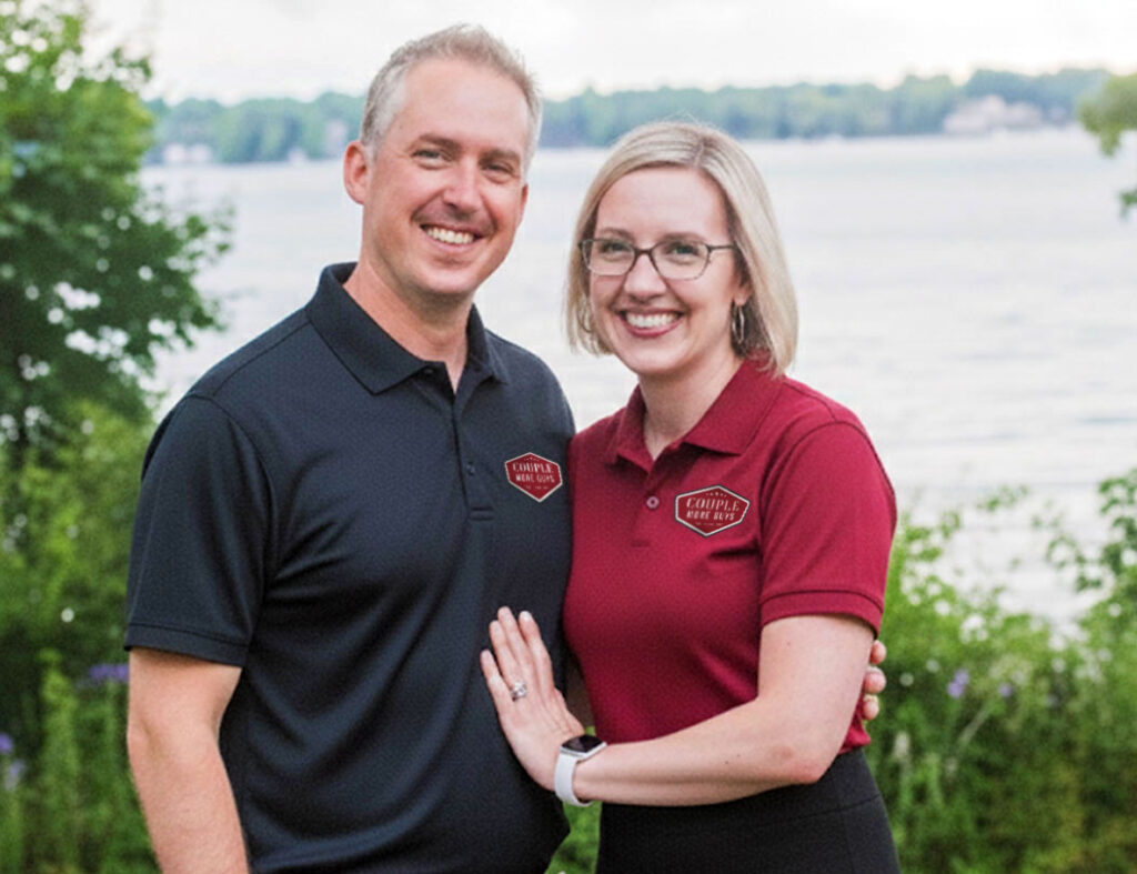 A smiling man and woman stand outdoors by a lake, wearing matching embroidered polos with Chaffee Real Estate logos. The man wears black and the woman wears red. Trees and water are visible in the background.
