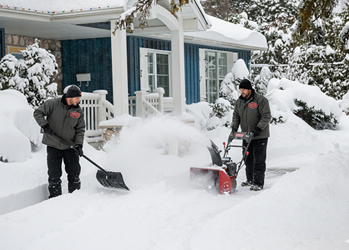 Two people in winter gear clear snow from a walkway; one uses a snow blower while the other shovels. Snow blankets the ground and a house with a porch is visible in the background.