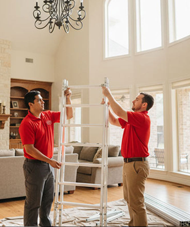 Two men in red polo shirts set up a ladder in a bright living room with large windows, a gray sofa, and a chandelier overhead. White cloths cover the wooden floor beneath them.