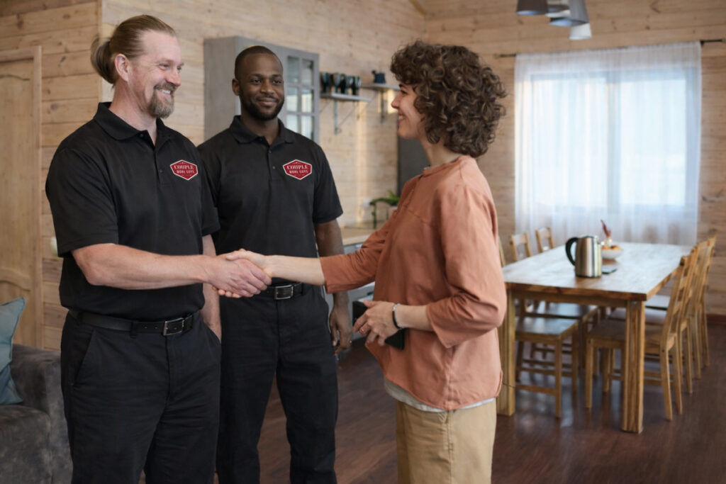A woman shakes hands with a smiling man in a black uniform, while another uniformed man stands beside them in a cozy, wooden dining area with a table and chairs.
