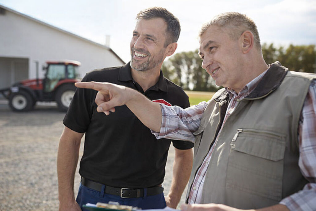 Two men stand outdoors near a tractor, one smiling in a black shirt and the other in a vest pointing into the distance, both appearing engaged in a friendly conversation on a farm.