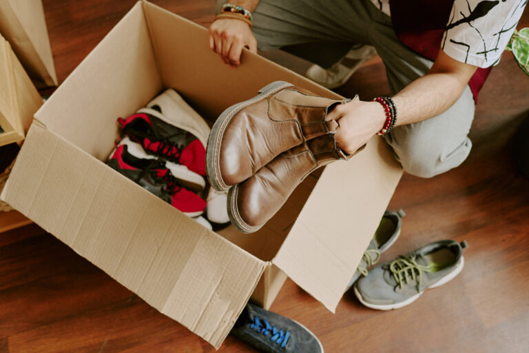 A person kneels on a wooden floor, holding brown boots above an open cardboard box filled with various shoes, including red sneakers. More shoes are scattered around the box.