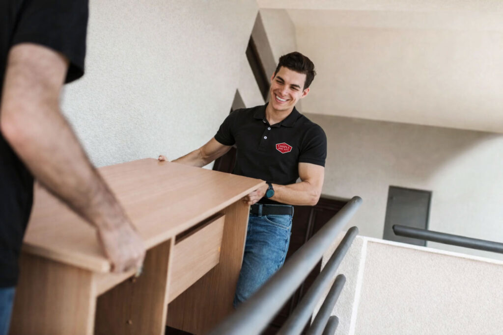 Two people carry a wooden desk up a staircase. One person, wearing a black polo shirt and jeans, is smiling as they lift the desk together. The setting appears to be inside an apartment building.