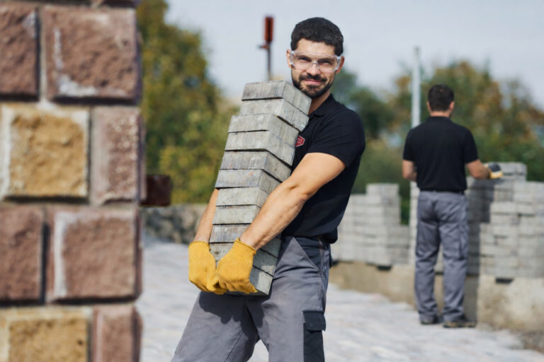 A man wearing safety glasses, yellow gloves, and work clothes carries a large stack of concrete bricks outside. Another worker stands in the background near more bricks and trees.