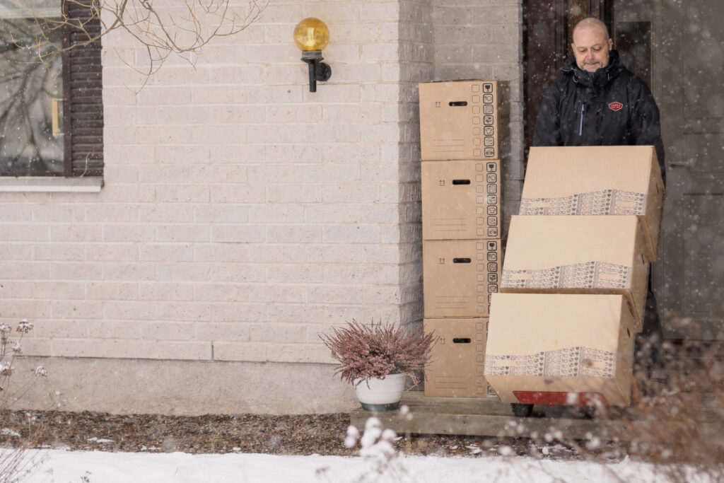 A person stands at the door of a house using a hand truck to move large cardboard boxes outside. Snow is falling, and a potted plant sits near the entrance.