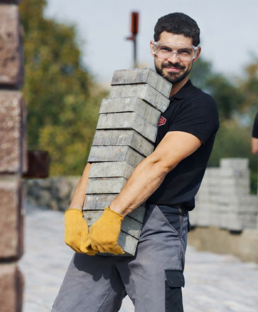 A man wearing safety glasses and yellow gloves smiles while carrying a tall stack of concrete pavers outdoors.