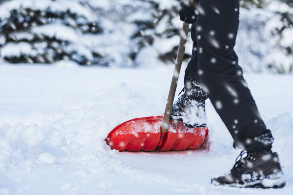 A person wearing black pants and boots is shoveling snow with a red snow shovel. Snow is falling, and trees covered in snow are visible in the background.
