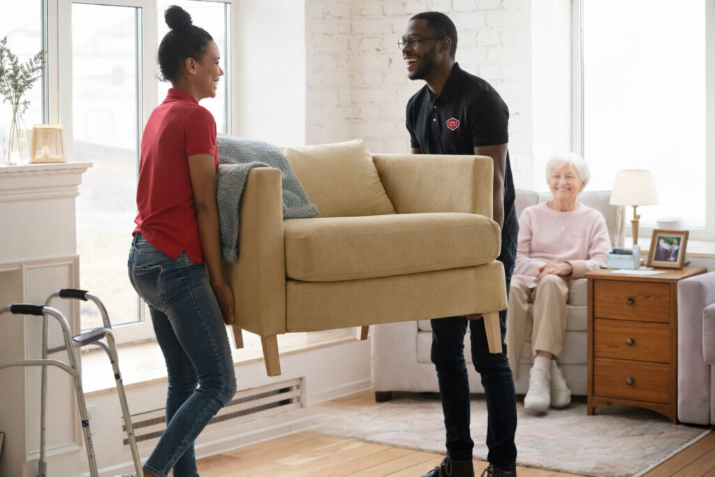 Two caregivers are smiling as they lift a yellow armchair in a bright living room. An elderly woman sits nearby, watching them, with a walker and a side table beside her.