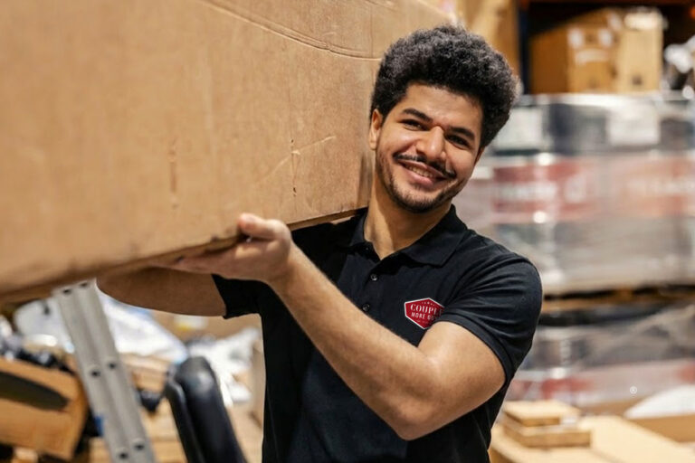 A smiling man in a black polo shirt carries a large cardboard box on his shoulder in a warehouse filled with boxes and supplies.