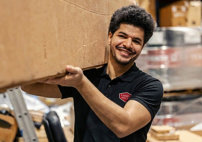 A smiling man in a black polo shirt carries a large cardboard box on his shoulder inside a warehouse filled with various packages and supplies.