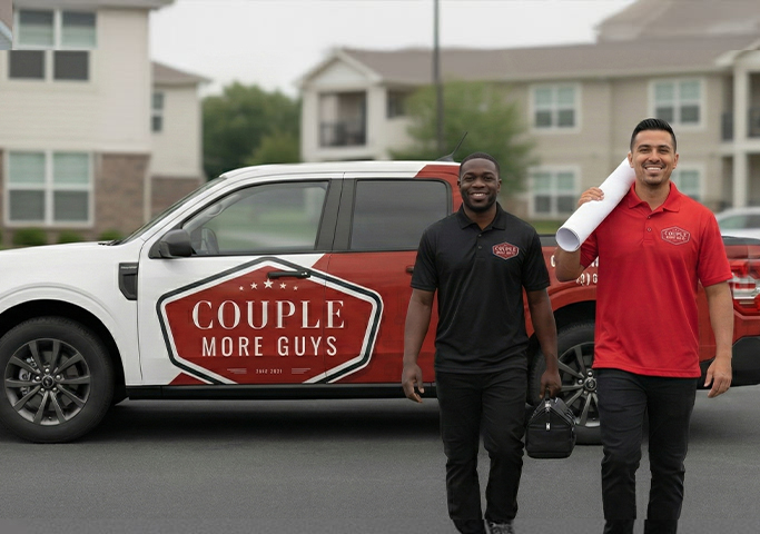 Two smiling men, one in a black polo and one in a red polo, walk in front of a branded pickup truck that reads “Couple More Guys” in a residential area, carrying tools and a rolled-up blueprint.