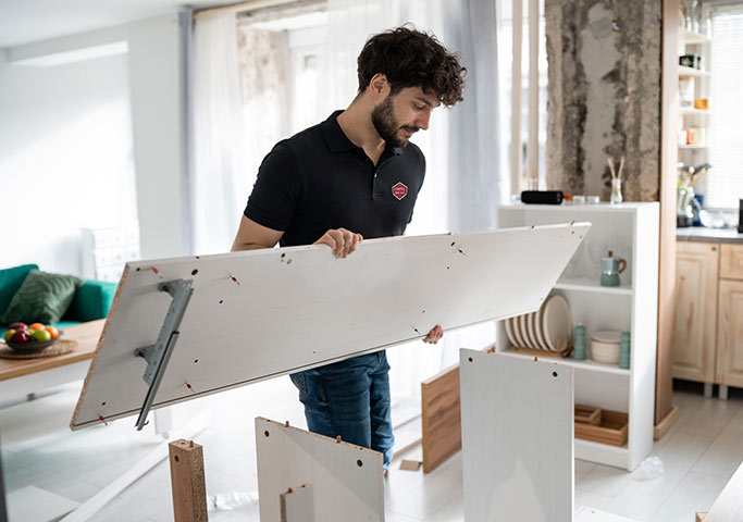 A man is assembling furniture in a bright room, holding a large white wooden panel and examining it, with other pieces and tools scattered around. Shelves and kitchen items are visible in the background.