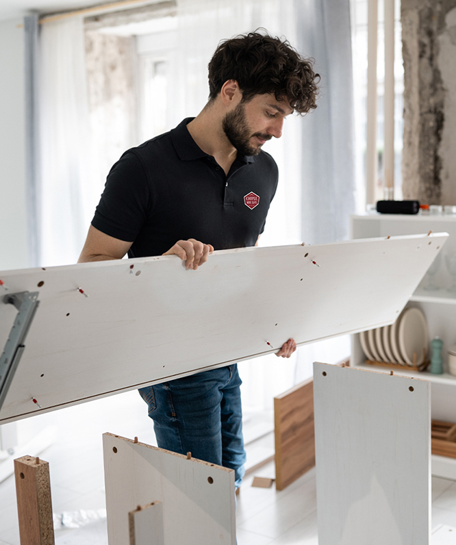 A man in a black polo shirt is assembling furniture indoors, holding a large white wooden board and standing beside other wooden panels in a bright room.