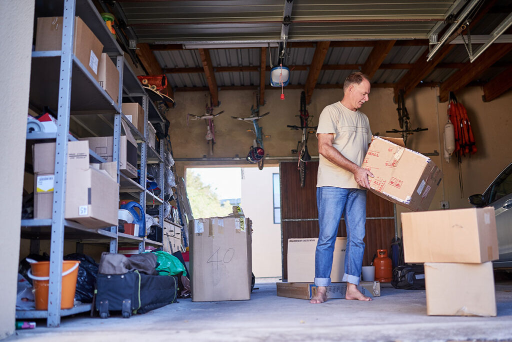A man stands barefoot in a garage, holding a cardboard box. Shelves filled with boxes and various items line the wall, and more boxes are scattered on the floor around him. A car is partially visible on the right.
