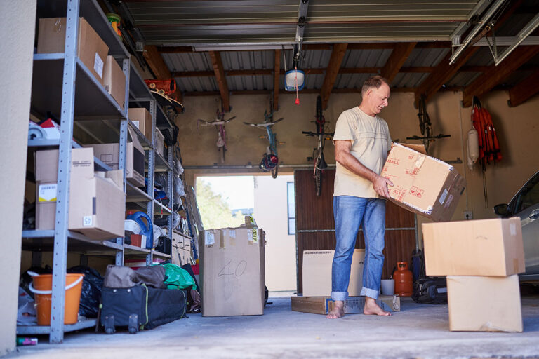 A man stands barefoot in a garage, holding a cardboard box. Shelves filled with boxes and various items line the wall, and more boxes are scattered on the floor around him. A car is partially visible on the right.