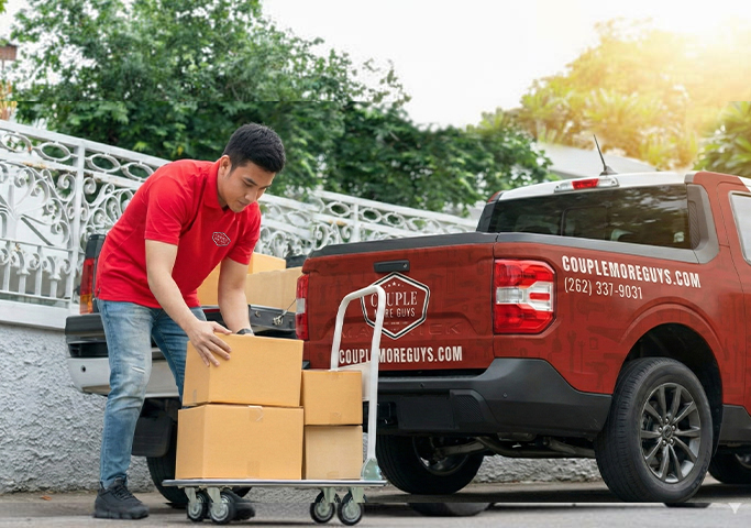 A man in a red shirt loads cardboard boxes onto a cart behind a red pickup truck branded with couplemoreguys.com and contact information, parked near a white fence on a sunny day.