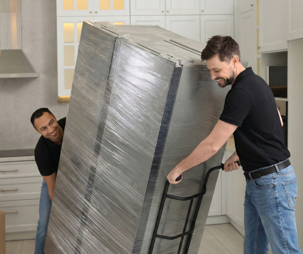 Two men in casual clothes carefully move a large, wrapped refrigerator on a hand truck in a modern kitchen with white cabinets.