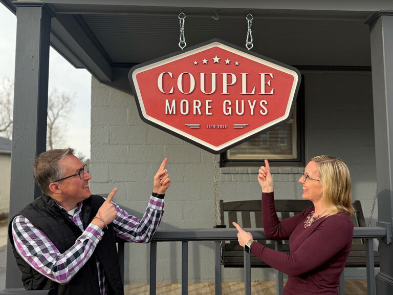 A man and a woman stand on a porch, smiling and pointing up at a large red sign that reads COUPLE MORE GUYS in white letters. The sign is hanging from the ceiling above them.