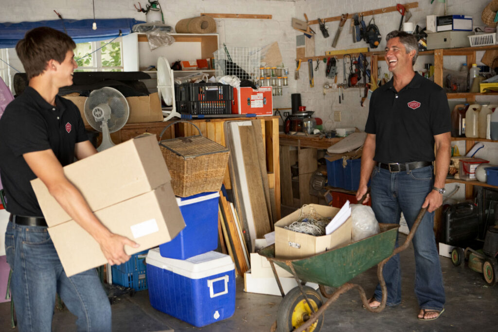 Two men in black polo shirts smiling while organizing a cluttered garage; one carries cardboard boxes and the other stands with a green wheelbarrow filled with miscellaneous items.