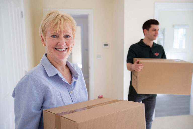 A smiling older woman and a younger man carry cardboard boxes inside a home, suggesting they are moving in or out. The woman is in the foreground, while the man is slightly blurred in the background.