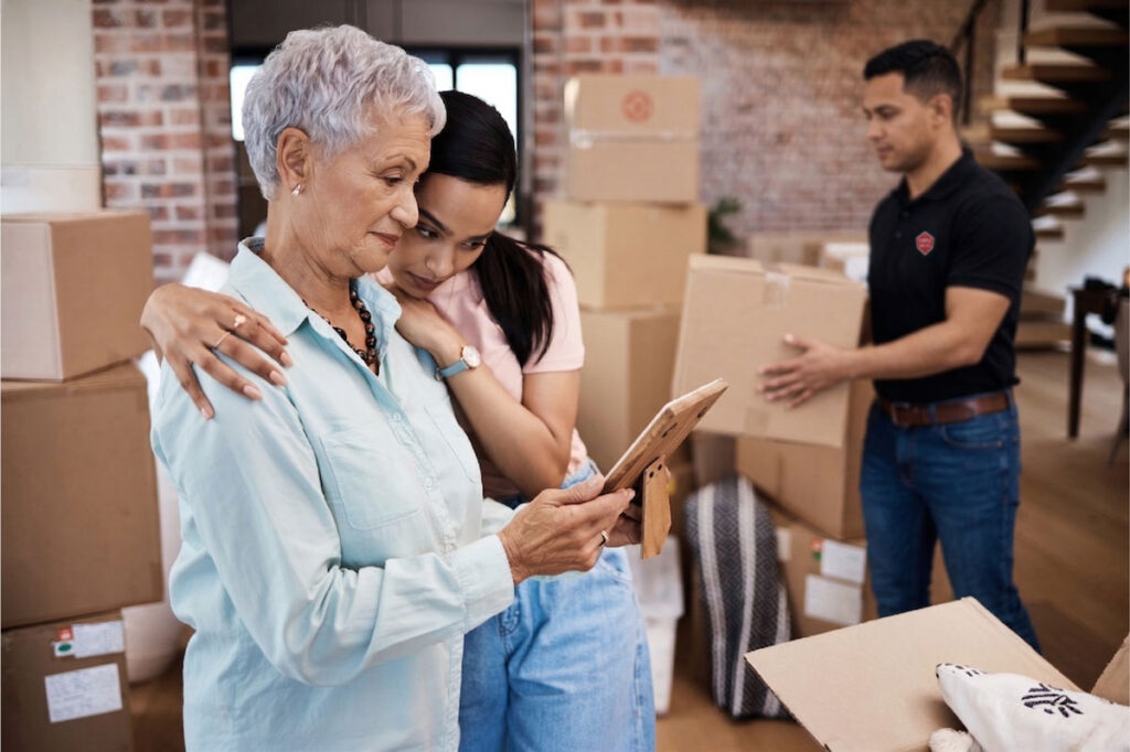An elderly woman and a young woman embrace while looking at a photo frame, surrounded by moving boxes in a home. A man in the background carries a box, suggesting they are moving or unpacking.