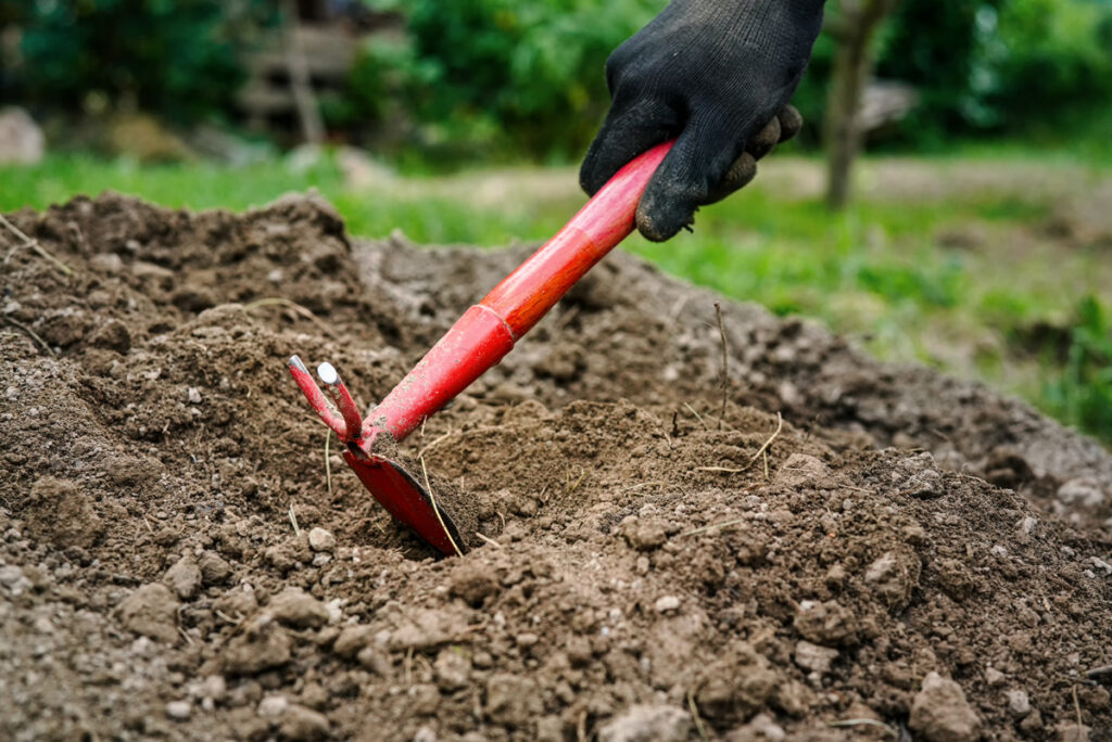 A person wearing a black glove uses a red hoe to till soil in a garden, with green plants and blurred foliage visible in the background.
