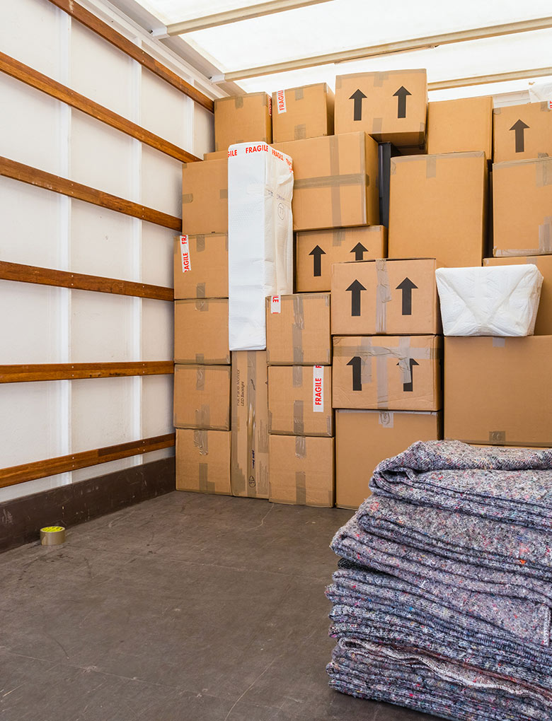 A moving truck interior with neatly stacked cardboard boxes, some labeled with arrows and fragile stickers. Folded moving blankets are on the floor in the foreground.