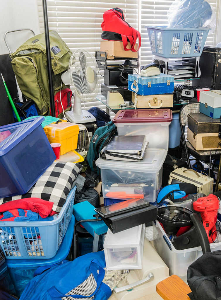 A cluttered room filled with various plastic bins, baskets, boxes, clothes, bags, a fan, and household items stacked and scattered, creating a chaotic and disorganized scene in front of a window with closed blinds.