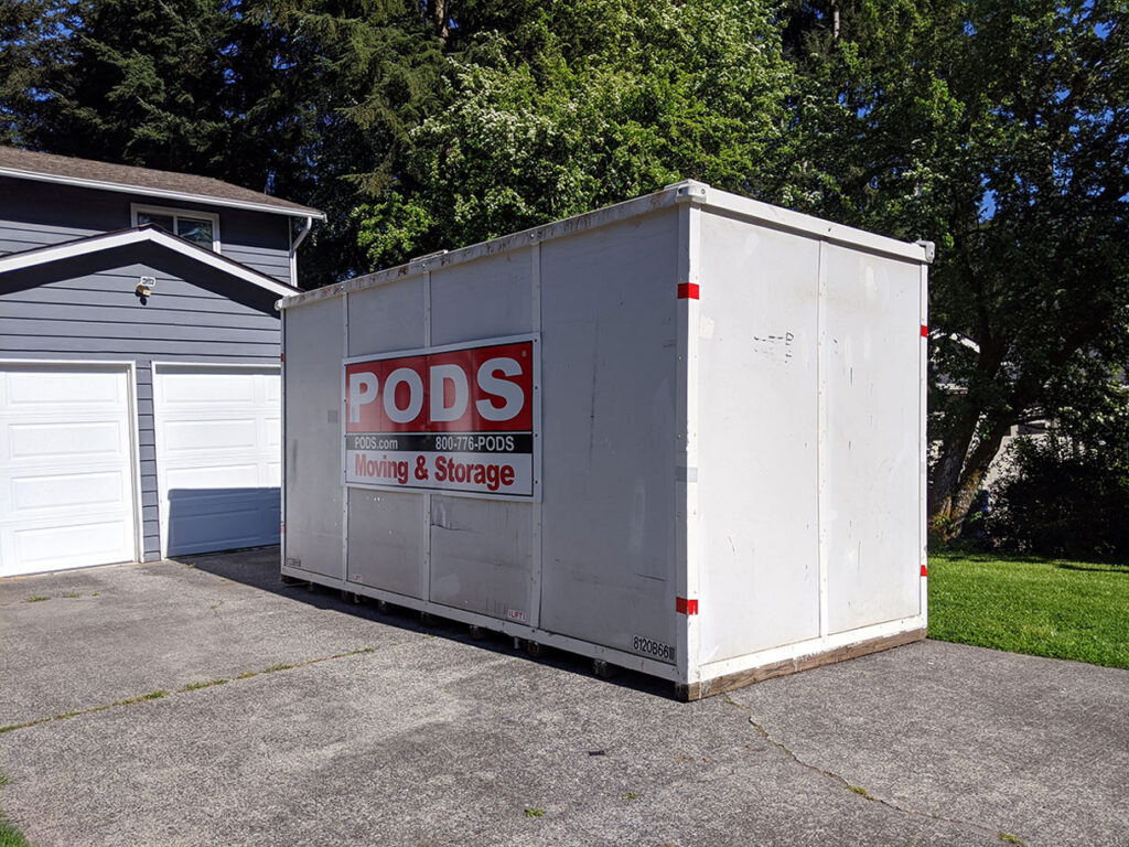 A large white PODS moving and storage container sits on a driveway in front of a gray house with white trim, next to a green lawn and tall trees in the background.
