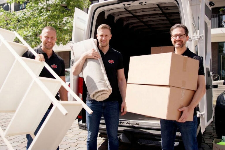 Three men smiling while unloading a van, carrying moving boxes, a rolled-up rug, and a white shelving unit on a sunny day. They appear to be working for a moving company.