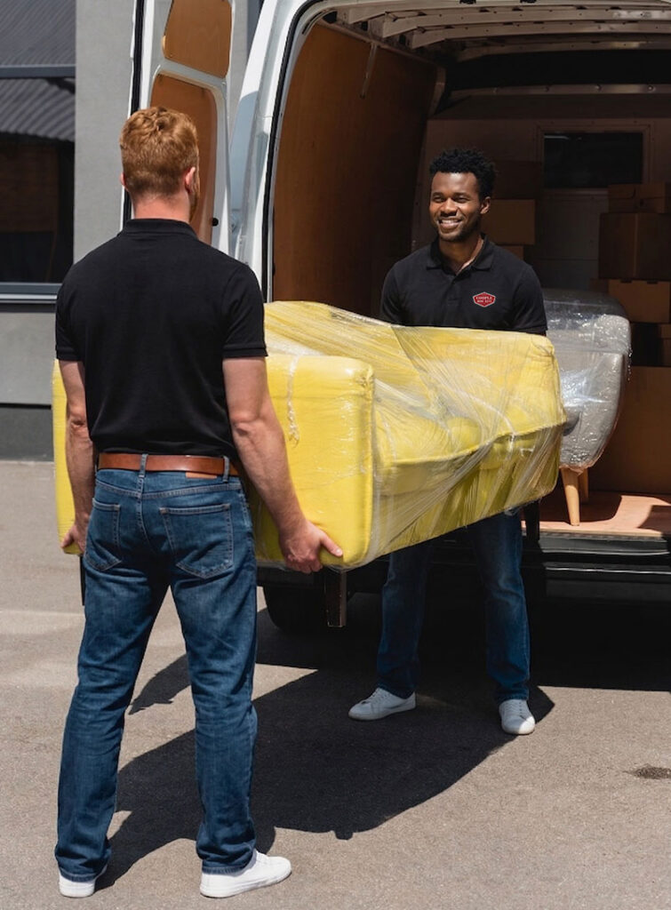 Two men, both wearing black shirts and jeans, are lifting a yellow couch wrapped in plastic and carrying it out of a white moving van. They appear to be moving furniture on a sunny day.