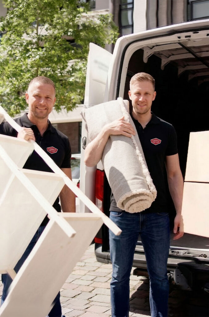 Two men in matching black shirts are unloading furniture and a rolled-up rug from a white van onto a sunny street with trees and buildings in the background.
