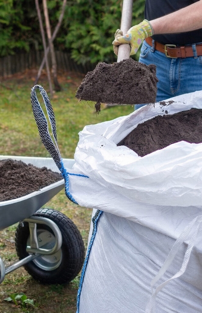 A person wearing gloves shovels dark soil from a large white bag, with a wheelbarrow filled with soil nearby in a garden setting.