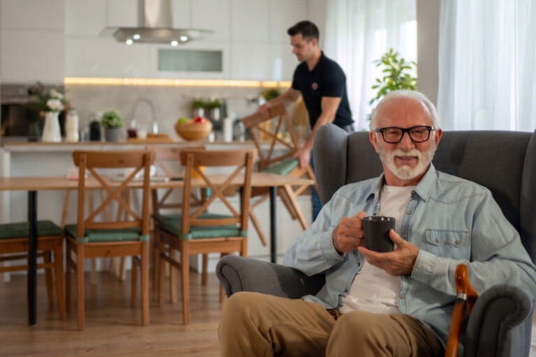 An older man with white hair and glasses sits in a gray armchair holding a mug, smiling. In the background, a younger man stands in a modern kitchen near a dining table.