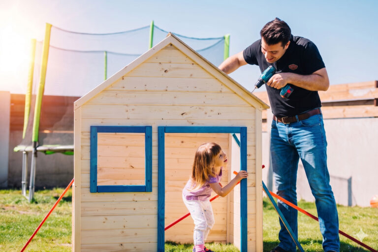 A man uses a power drill to build a small wooden playhouse in a backyard while a young girl smiles and looks up at him from inside the structure. A trampoline is visible in the background.