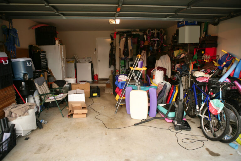 A cluttered garage filled with various items including bicycles, boxes, pool noodles, laundry machines, storage bins, household items, and tools, with little open floor space visible.