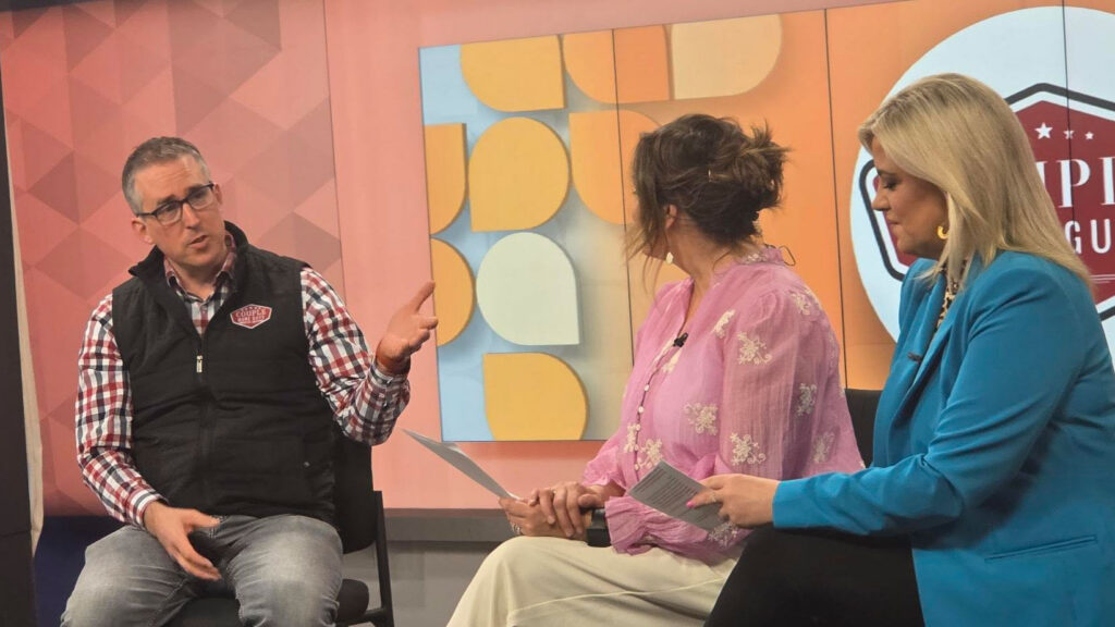Three people sit and talk in a TV studio. The man on the left gestures while speaking, wearing glasses and a checkered shirt with a black vest. Two women face him, holding papers. The colorful set displays geometric shapes and a logo.
