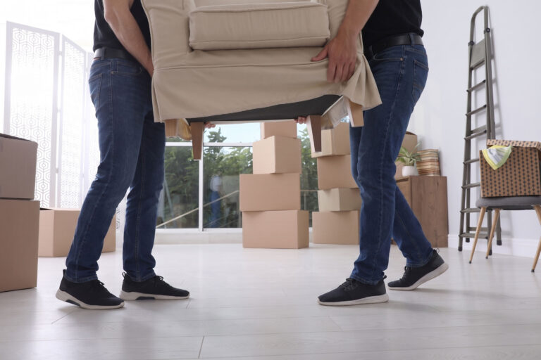 Two people in jeans and black shirts lift a beige armchair in a bright room filled with packed cardboard boxes. A ladder, baskets, and a large window are visible in the background, suggesting a home move.
