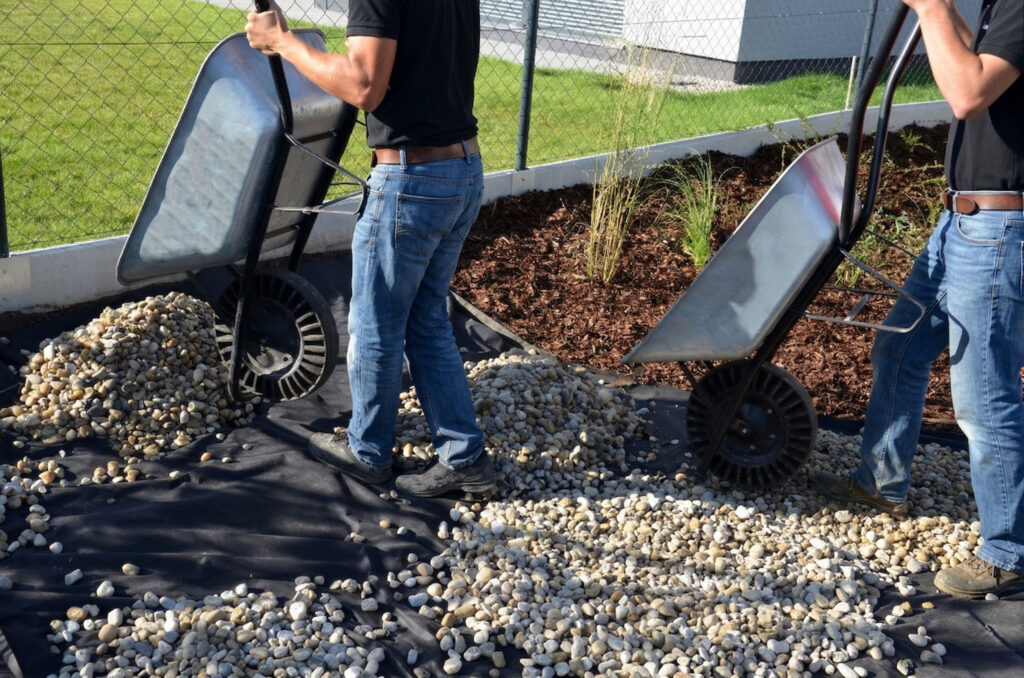 Two people in jeans and black shirts dump rocks from wheelbarrows onto a garden bed covered with landscaping fabric, near a fence and mulched plants.
