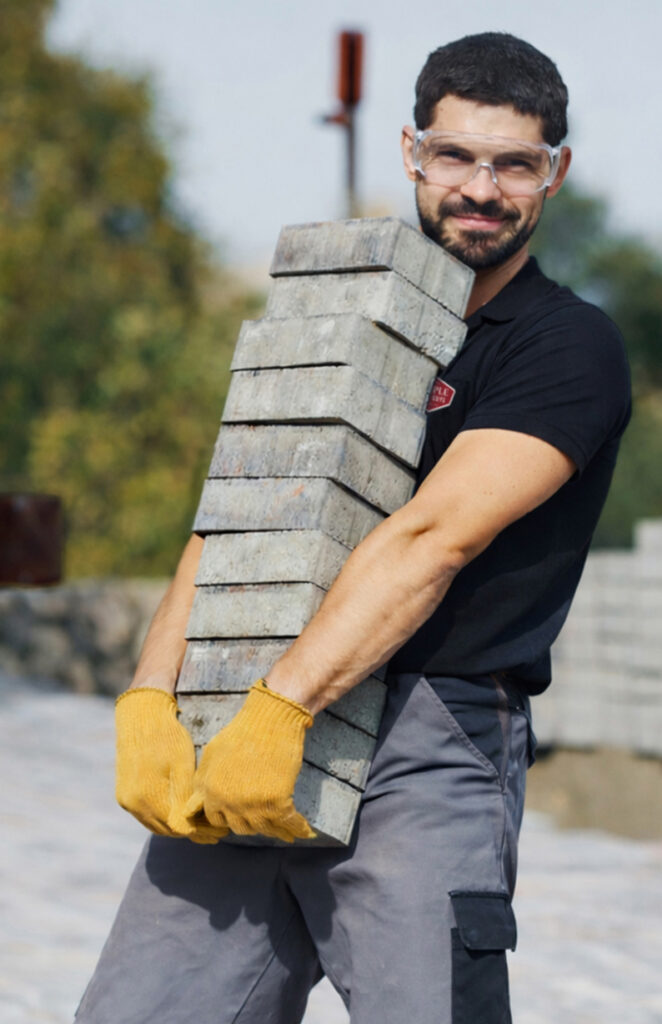 A man wearing safety glasses and yellow gloves carries a tall stack of concrete paving blocks outdoors, smiling at the camera.