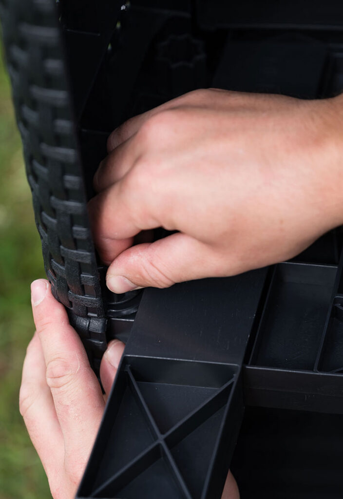 Close-up of two hands assembling a black plastic or wicker furniture piece outdoors, fitting a panel into place.