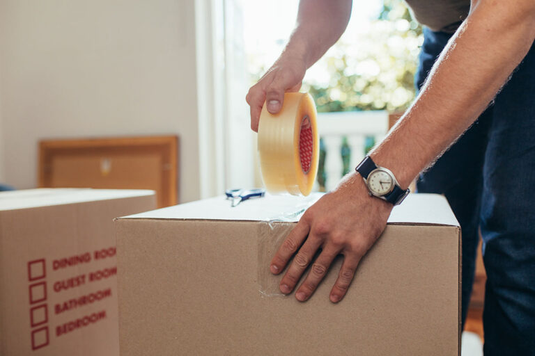 A person seals a cardboard moving box with clear packing tape. A checklist with rooms is printed on the box. The person wears a wristwatch and sunlight streams through a window in the background.