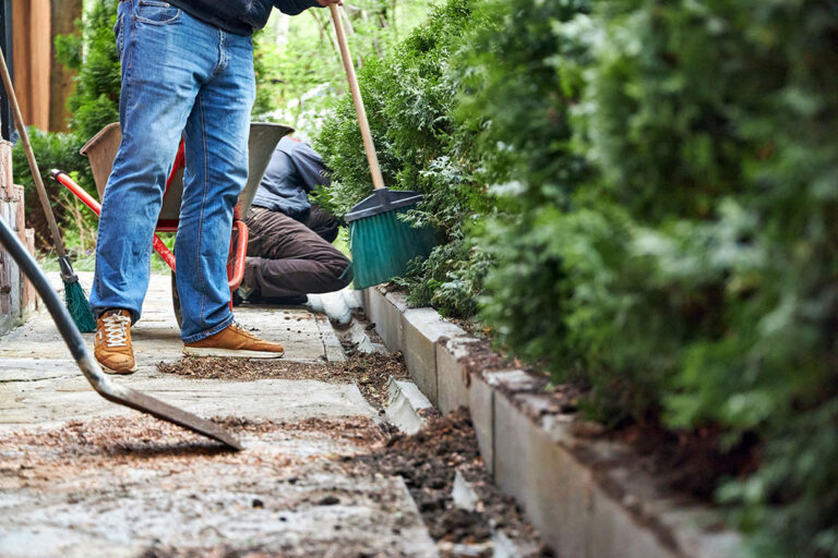 Two people working outdoors on a garden path; one sweeps near the edge with a broom while the other kneels, tending to plants or performing maintenance tasks among green bushes.