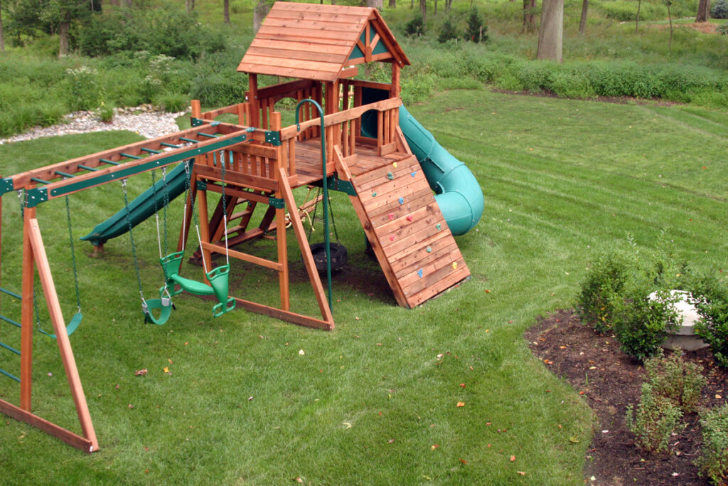 A wooden backyard playground set with swings, a slide, a climbing wall, and a small playhouse stands on a green lawn, surrounded by trees and garden beds.
