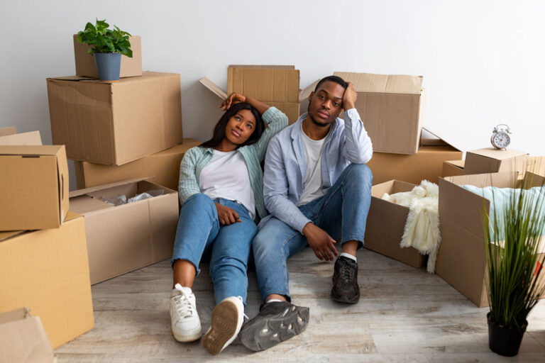 A couple sits on the floor, looking tired among stacked cardboard boxes and household items, suggesting they are in the process of moving into a new home.