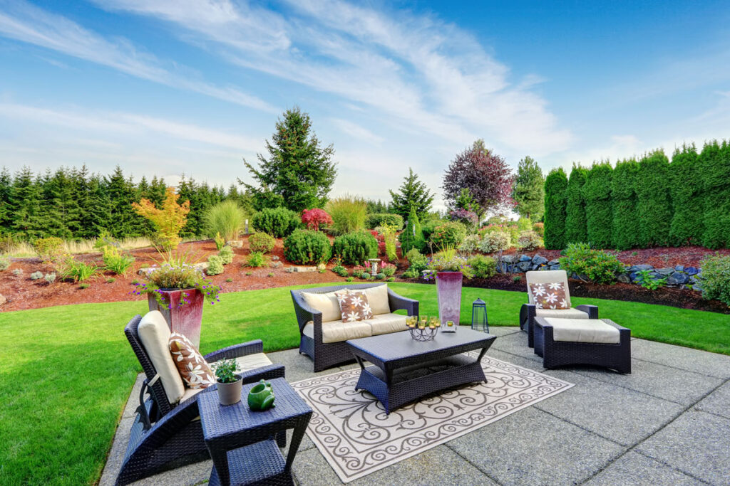 A patio with cushioned wicker furniture and a decorative rug overlooks a landscaped backyard with green grass, colorful shrubs, trees, and a clear blue sky.