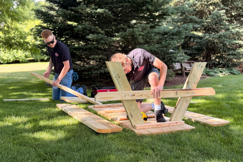 Two people are assembling a wooden structure on grass in a sunny outdoor setting, with one person kneeling to connect beams and the other holding planks. Trees and tools are visible in the background.