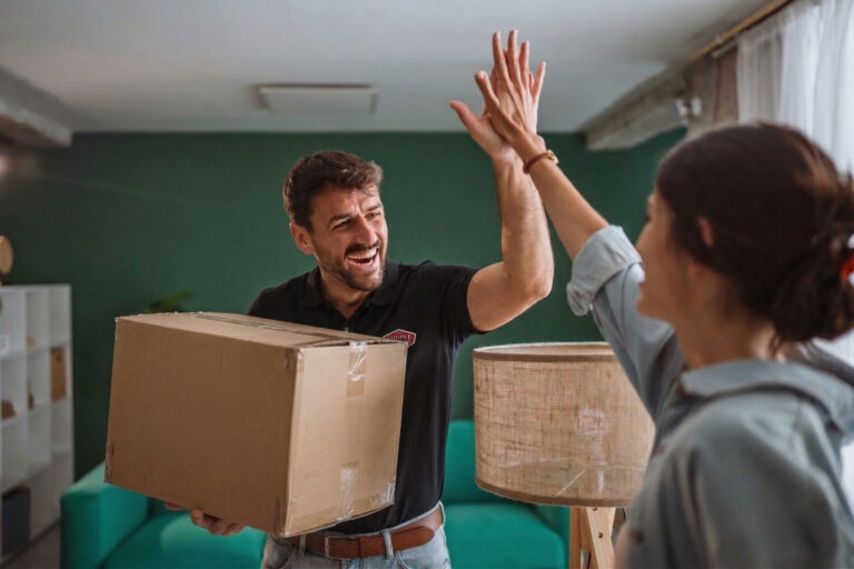 A man holding a cardboard box smiles and gives a high-five to a woman inside a brightly lit room with green walls and a teal sofa in the background.